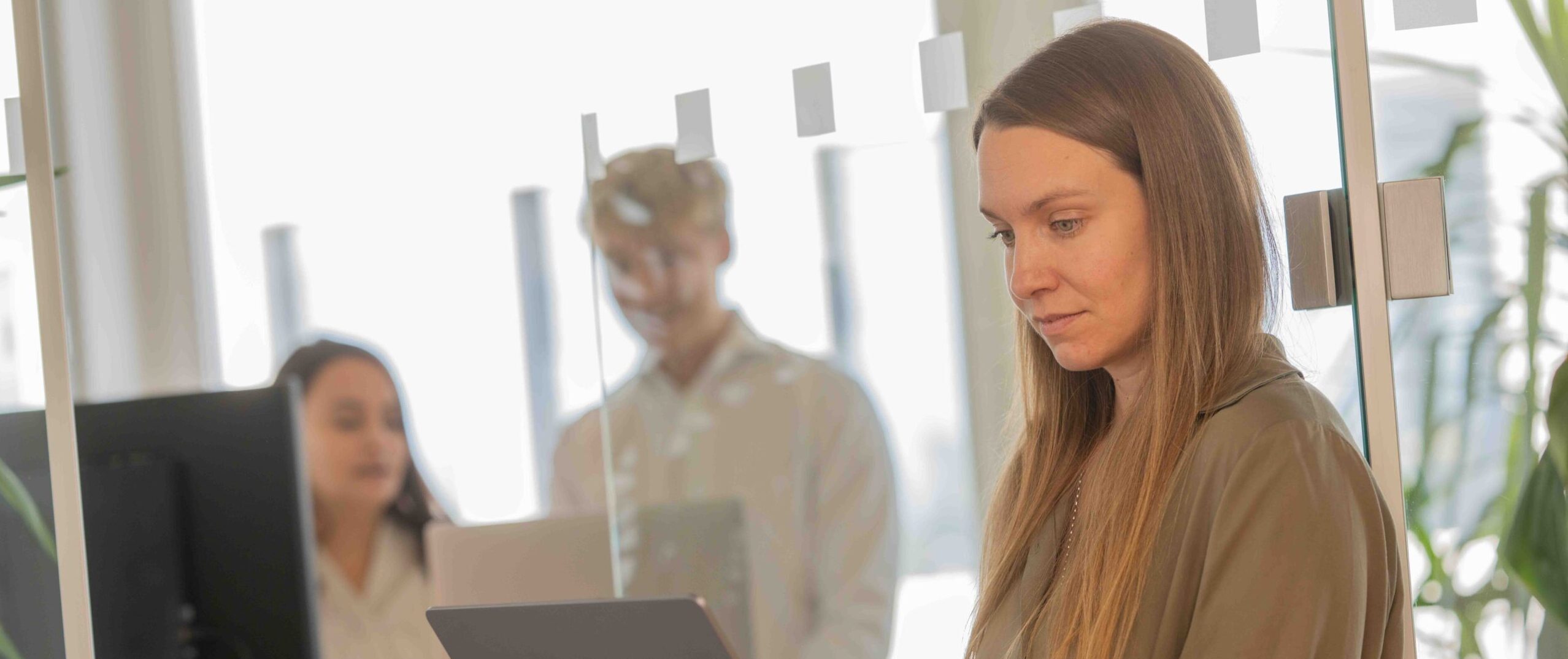 Alena schaut auf ihren Laptop, w&auml;hrend Melanie und Sandro im Hintergrund in einer Besprechung sind.